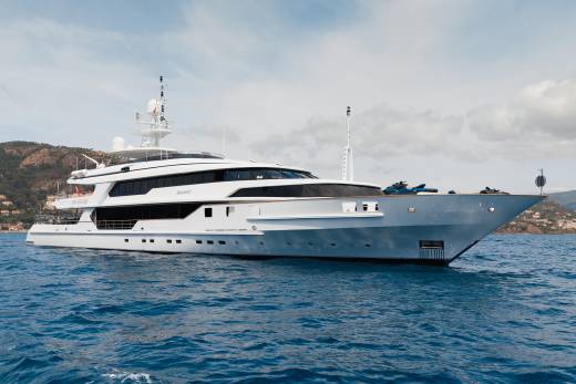 A large white yacht floats on blue water in the foreground, with a hilly, tree-covered coastline under a cloudy sky in the background.