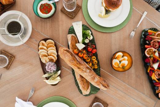 A wooden table is laden with a variety of appetizers, including bread, cheese, dips, and fruit platters, with a blurred background suggesting an outdoor setting.