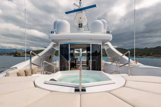 A luxurious yacht with a jacuzzi in the foreground sails on the sea with a cloudy sky and distant hills in the background.