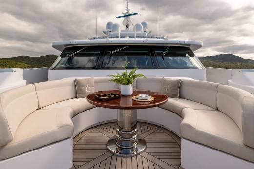 A circular outdoor seating area with a table and plant is on the deck of a yacht, with a cloudy sky and hills in the background.