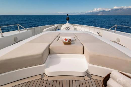 A luxurious yacht's bow with plush seating in the foreground faces a calm blue sea and distant, hazy mountains under a clear sky.
