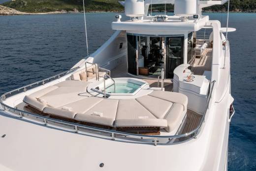 A luxurious yacht with a large sunbathing area and jacuzzi in the foreground sails on calm blue water with islands in the background.