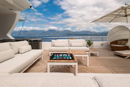 White sofas and a backgammon board are on a wooden deck in the foreground, with a serene ocean and distant mountains under a blue sky in the background.