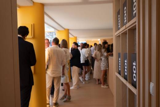 A man in a suit stands in the foreground to the left, while a crowd of people mingle in the background of a brightly lit, yellow-accented room with a shelving unit on the right.