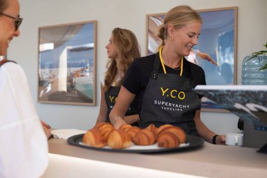 A smiling woman in a Y.CO apron serves coffee to a customer in the foreground, with another woman and framed artwork visible in the background.