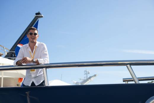 A man in a white shirt smiles on a yacht's deck with a clear blue sky and another boat in the background.