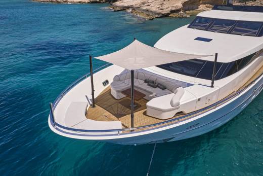 A luxurious white yacht with a shaded seating area in the foreground floats on clear turquoise water near a rocky, tree-lined coastline in the background.