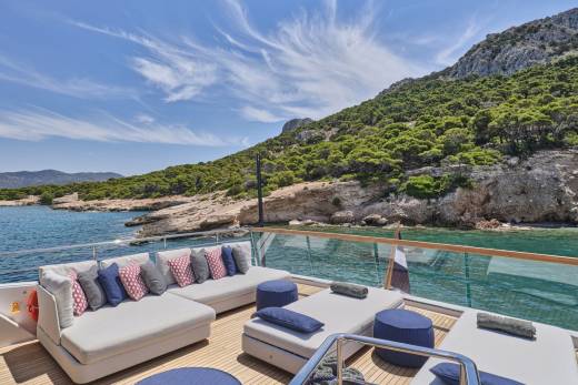 A luxurious yacht deck with plush seating in the foreground overlooks a serene bay with a lush, green mountainside in the background.