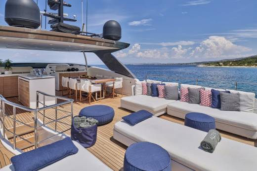 A luxurious yacht deck with white lounge chairs and blue cushions in the foreground is set against a backdrop of the sparkling blue ocean and a distant, tree-covered coastline under a clear sky.