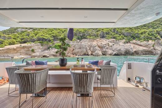 Wicker chairs surround a wooden table on a yacht's deck, with a lush, rocky coastline visible in the background.