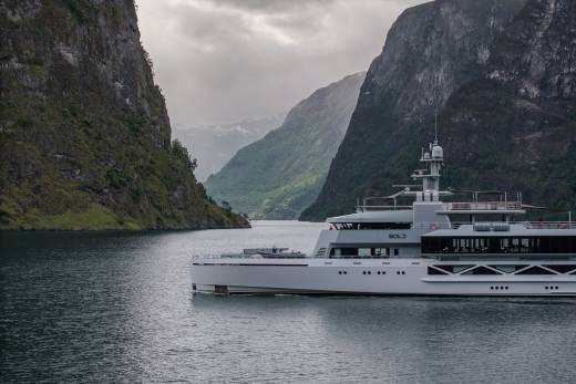 A sleek, modern yacht sails on calm water between towering, verdant cliffs under a cloudy sky.