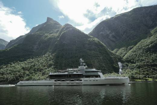 A large white yacht sails on calm water in the foreground, with lush green mountains and a waterfall in the background.
