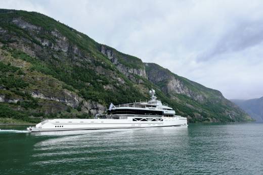 A large white yacht cruises on calm water in the foreground, with steep, green, tree-covered mountains rising dramatically in the background.