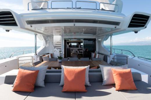 A luxurious yacht's outdoor seating area with orange cushions in the foreground is set against a backdrop of the calm ocean and distant mountains under a clear sky.