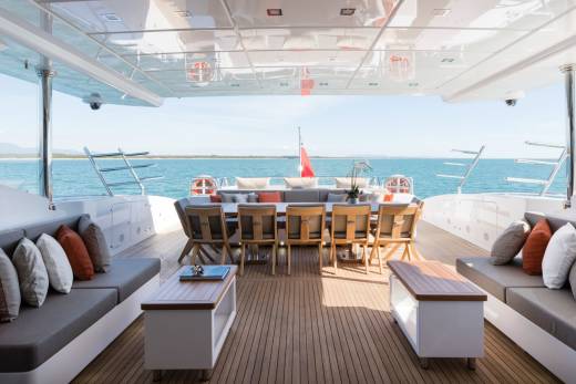 A luxurious yacht deck with comfortable seating and a dining area overlooks a calm blue sea under a clear sky.