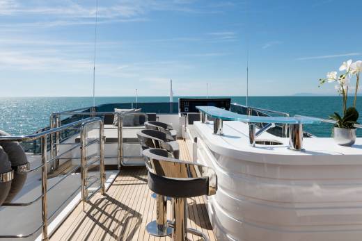 A white bar with silver stools sits on a wooden deck in the foreground, with the calm blue ocean and clear sky stretching to the horizon in the background.