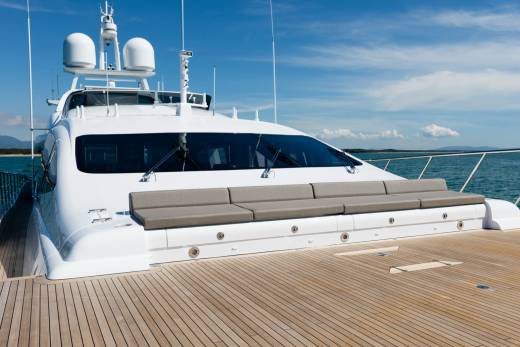 A luxurious white yacht with a spacious wooden deck in the foreground is anchored in calm blue waters with a distant coastline under a clear sky.