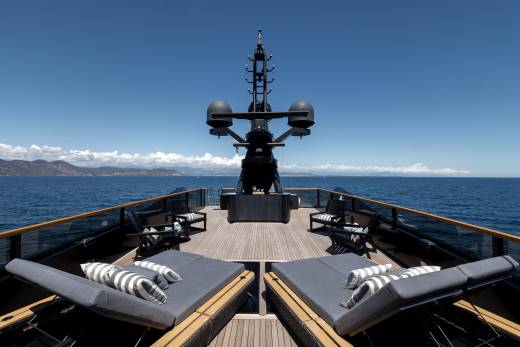 Two plush lounge chairs with striped cushions sit on a wooden deck in the foreground, while a large, dark radar array dominates the background against a clear blue sky and distant coastline.