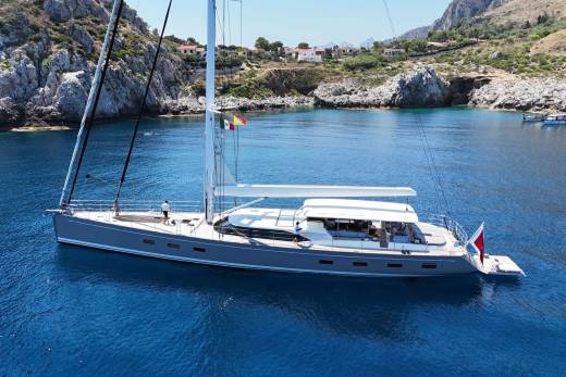 A sleek gray sailboat with white sails glides through calm blue water in a secluded cove with rocky, tree-covered hills rising in the background.
