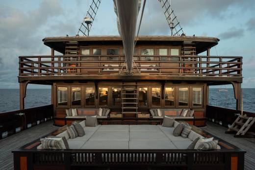 A spacious, cushioned deck with seating in the foreground leads to a multi-level wooden boat with numerous windows and railings, set against a calm ocean and cloudy sky in the background.