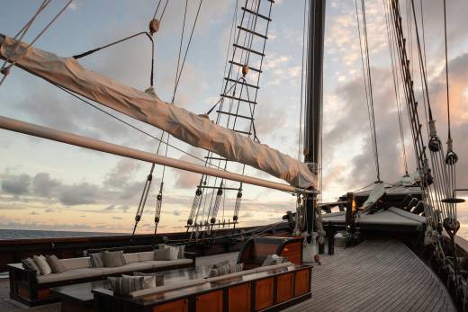 A luxurious wooden deck with plush seating in the foreground is set against a dramatic sky with clouds and the rigging of a sailing ship in the background.