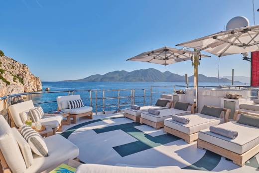 Sun loungers and seating areas on a yacht's deck overlook a calm sea and distant, mountainous coastline under a clear blue sky.