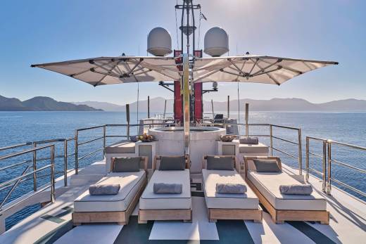 Four sun loungers with grey cushions are arranged on the deck of a yacht, with a clear blue sky and distant, hazy mountains in the background.