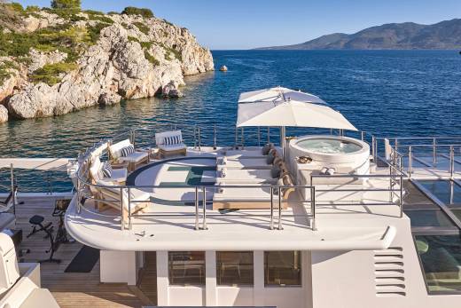 A luxurious yacht with a hot tub and seating area in the foreground is anchored in a calm bay with rocky cliffs and distant hills in the background.
