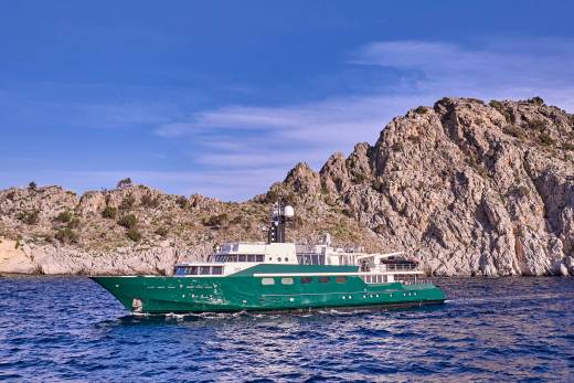 A green yacht sails on blue water in the foreground, with rugged, rocky cliffs and a clear blue sky in the background.