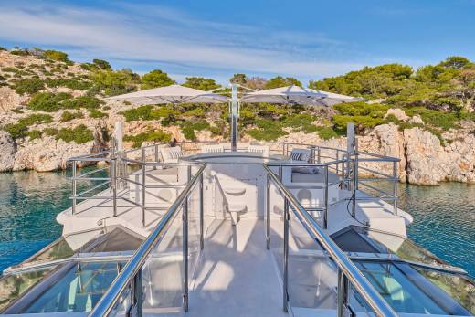 A luxurious yacht with open-air seating and umbrellas is anchored in a calm, turquoise bay surrounded by rocky, tree-covered hills under a clear blue sky.
