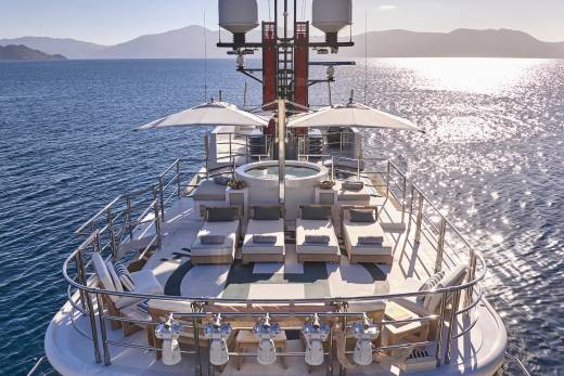 Sun loungers and a hot tub are arranged on the deck of a yacht in the foreground, with calm blue water and distant, hazy mountains in the background.