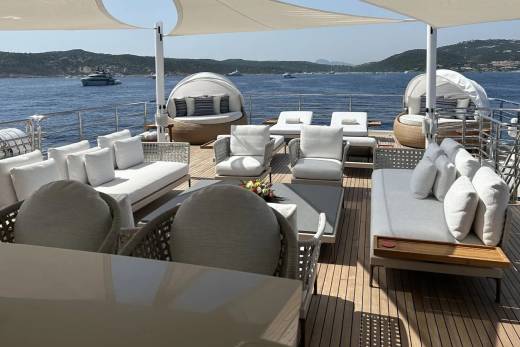Plush white outdoor furniture is arranged on a yacht's deck, with a calm blue sea and distant boats under a bright sky in the background.