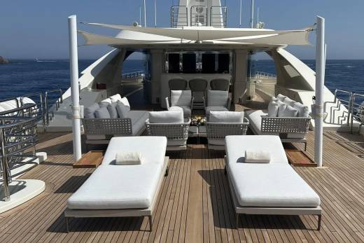 Two white lounge chairs with pillows sit on a wooden deck in the foreground, while a luxurious yacht with seating and awnings is visible in the background against a clear blue sky and ocean.