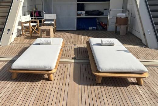 Two white cushioned lounge chairs with wooden frames sit on a teak deck in the foreground, while white chairs and a table are visible in the background.