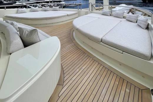 Plush, cream-colored sunbeds with pillows rest on a polished wooden deck in the foreground, while a harbor with docked boats is visible in the blurred background.