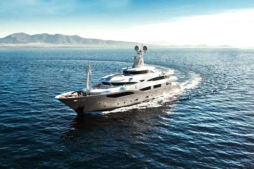 A large, white yacht cruises through calm blue water, leaving a foamy wake, with hazy mountains visible on the horizon under a bright sky.
