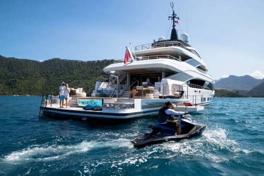 A person rides a jet ski in the foreground with a large yacht anchored in the background, surrounded by lush green hills under a clear blue sky.