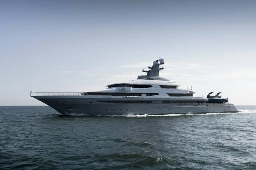 A sleek, gray superyacht cruises on calm, dark blue water under a pale, hazy sky.