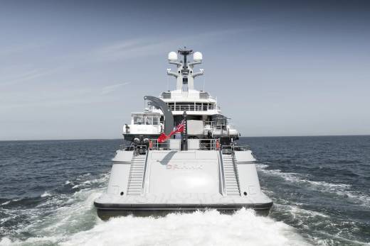 A large white yacht with a red flag sails through calm blue water, leaving a foamy wake under a clear sky.
