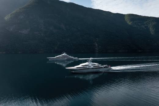 Two sleek yachts glide across a calm, dark blue body of water, leaving white wakes, with a majestic, tree-covered mountain range rising in the background under a hazy sky.