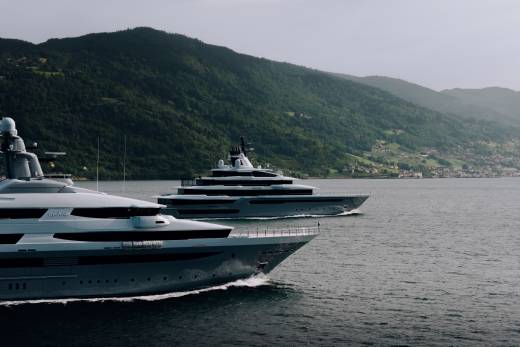Two large yachts sail on calm water in the foreground, with a lush green mountainside dotted with houses in the background.
