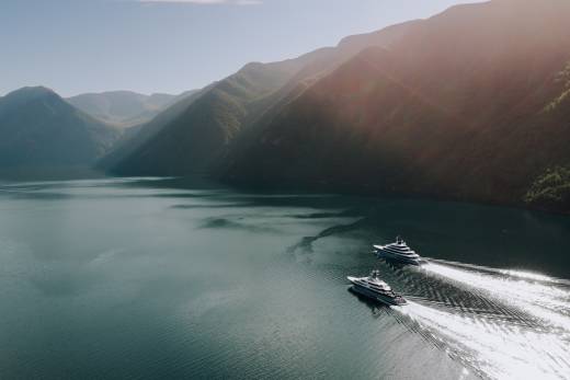 Two sleek yachts glide across a calm, expansive lake, leaving white wakes that stretch towards a backdrop of sun-drenched, tree-covered mountains.