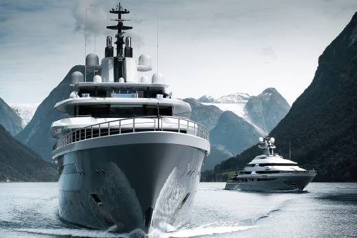 A large, modern yacht dominates the foreground, with another yacht trailing behind it in a serene, mountainous fjord.