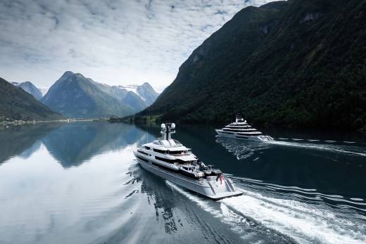 Two luxurious yachts cruise through a calm, reflective fjord, with dramatic, tree-covered mountains rising in the background under a cloudy sky.