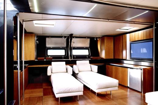 Two white chaise lounges sit on a polished wooden floor in the foreground, with a wall of cabinets, a television, and windows in the background.