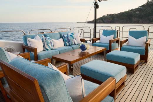 A luxurious outdoor seating area with light blue cushions and wooden frames is set on a yacht's deck, with the calm ocean and a distant, tree-covered coastline in the background.