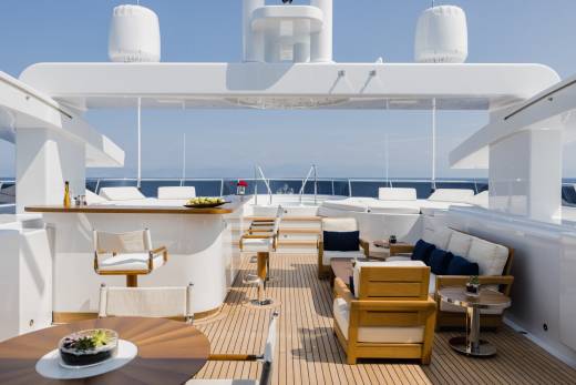 A luxurious yacht deck with a bar and seating area in the foreground overlooks a calm blue ocean under a clear sky.