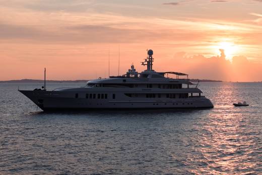 A large yacht floats on calm water in the foreground, with a small dinghy nearby, as the sun sets in the background, casting a warm glow across the sky.