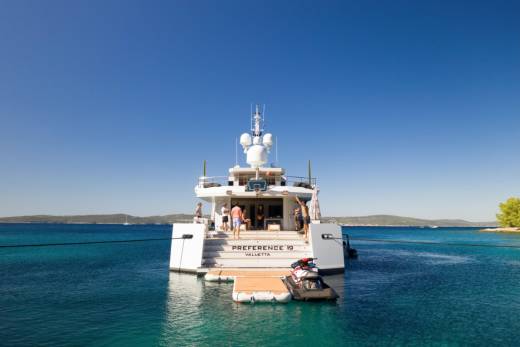 A large white yacht with people on its deck is anchored in clear turquoise water, with a jet ski attached to a floating platform, and a distant, tree-covered island under a bright blue sky.