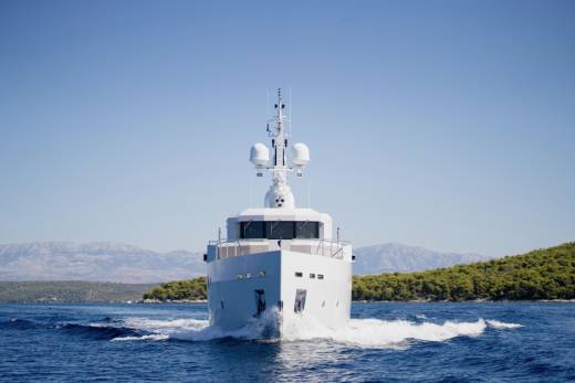 A white yacht cuts through blue water in the foreground, with a tree-covered island and hazy mountains in the background under a clear sky.